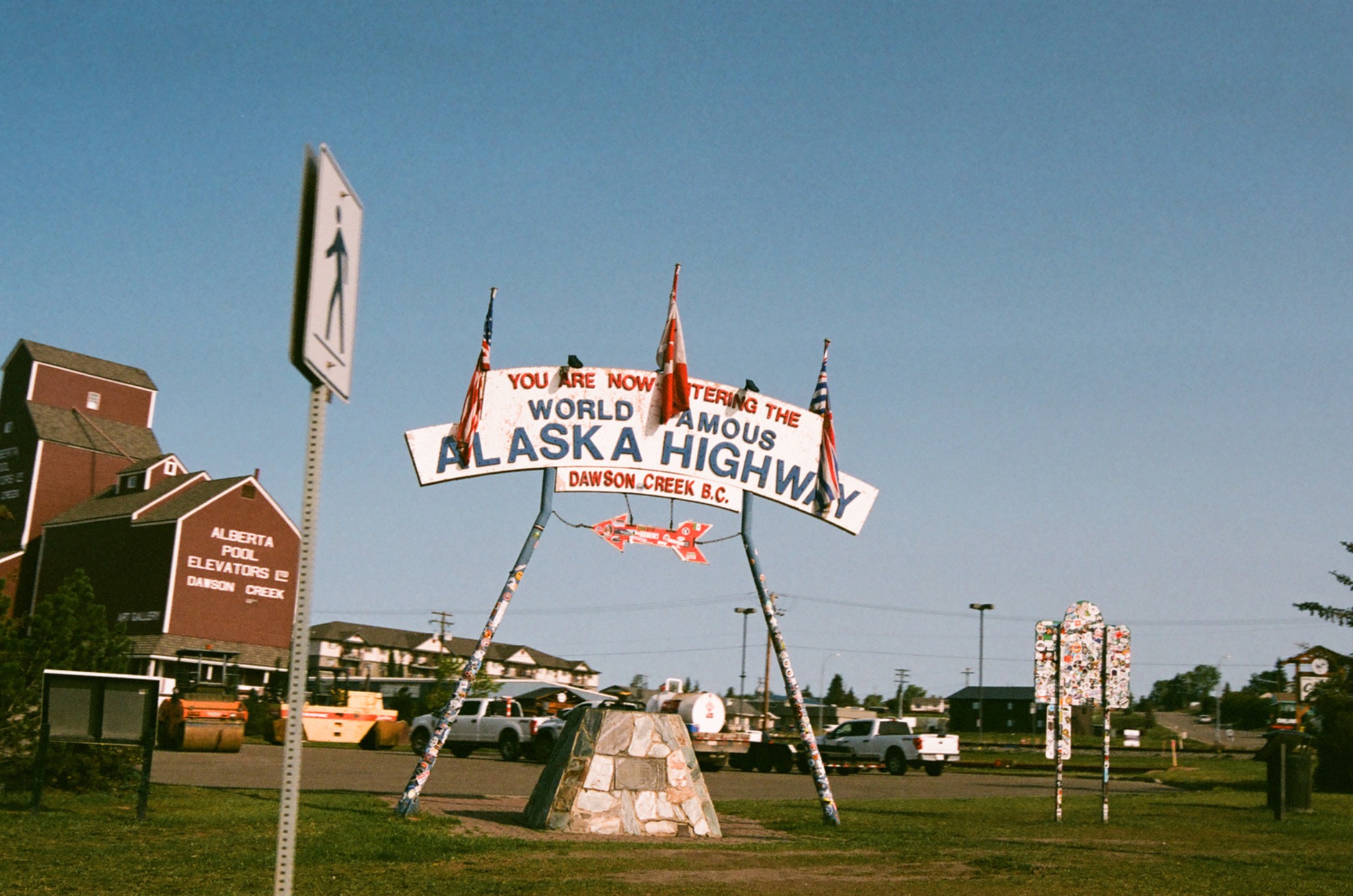 large sign for alaska highway in dawson creek with red buildings and vehicles