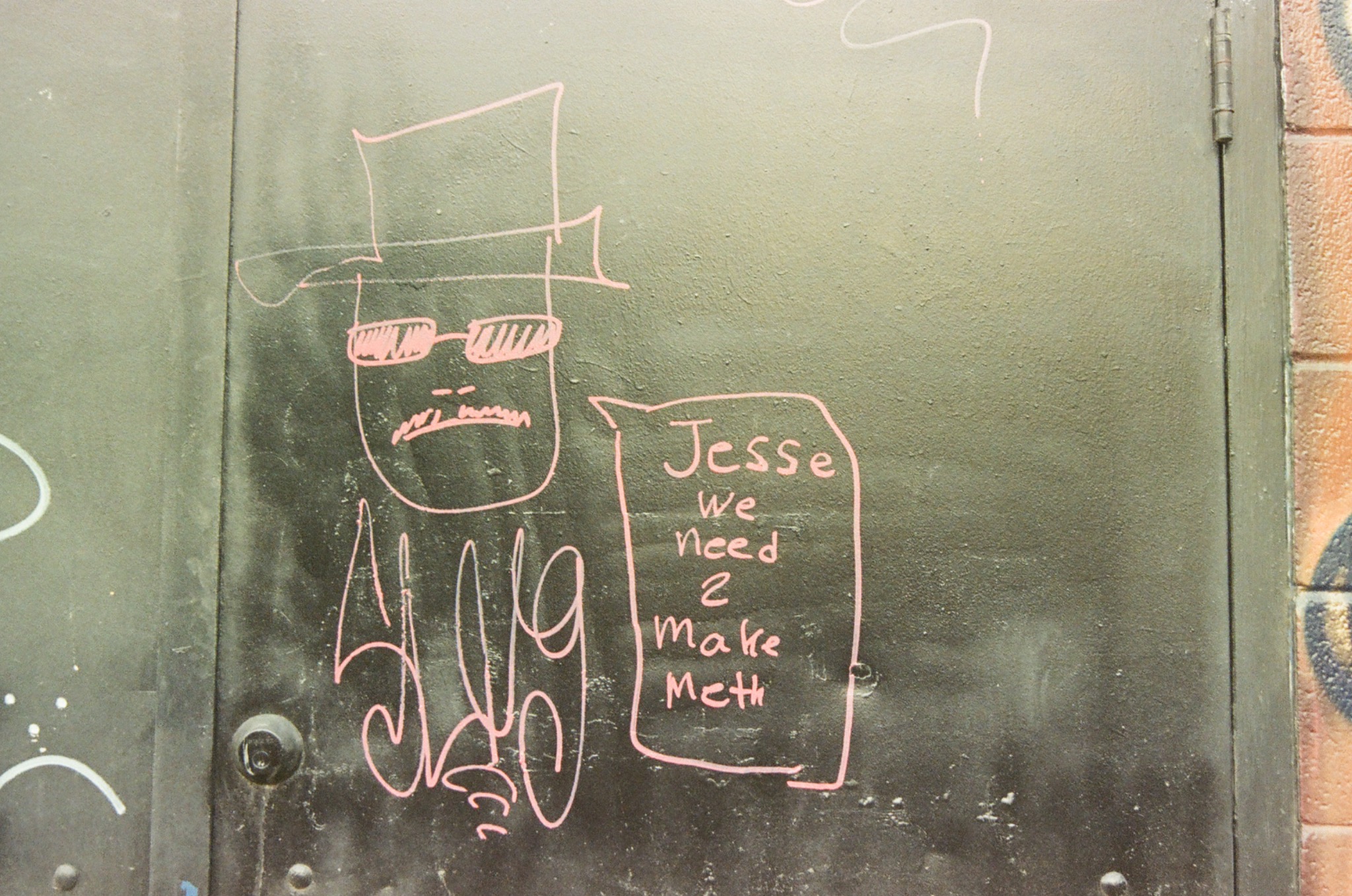 pink graffiti of a man in a hat and sunglasses with a speech bubble on a dark metal door.