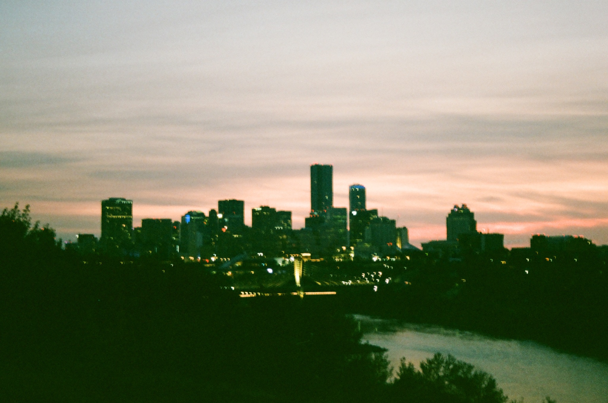 blurry cityscape at dusk with dark trees, a river, and illuminated buildings under a pale sky.