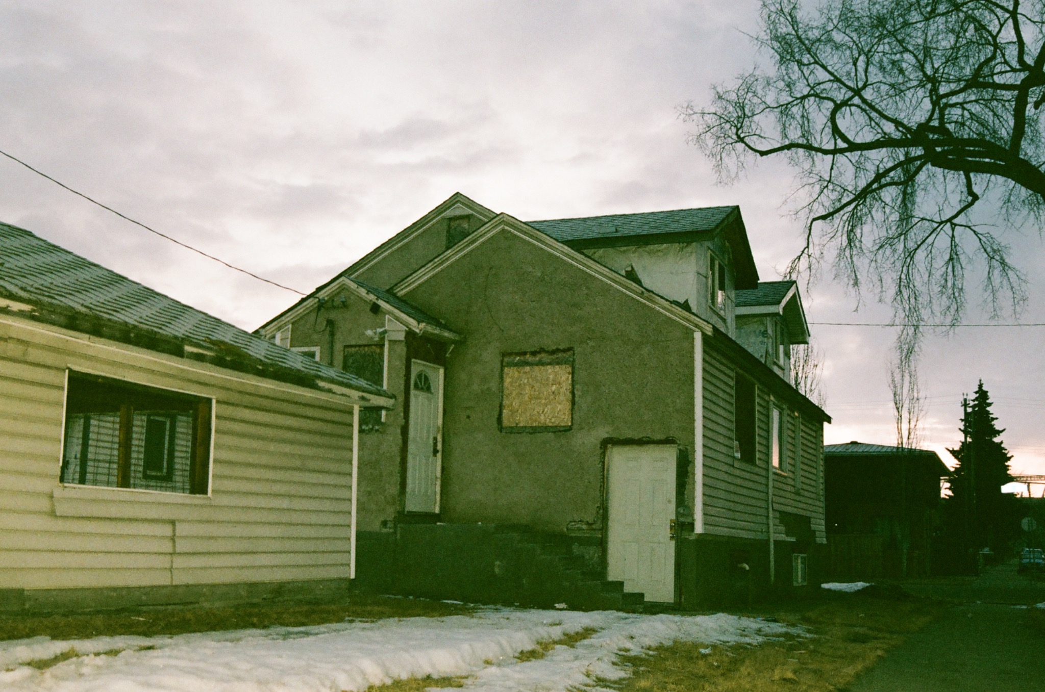 two weathered houses under an overcast sky with bare trees and patches of snow on the ground.