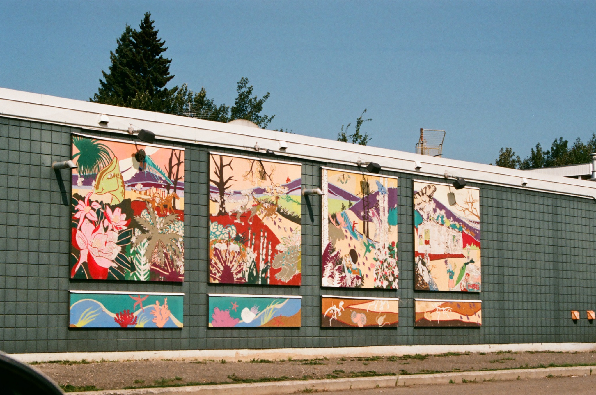 four colorful murals on a gray tiled building wall under a clear blue sky with green trees.