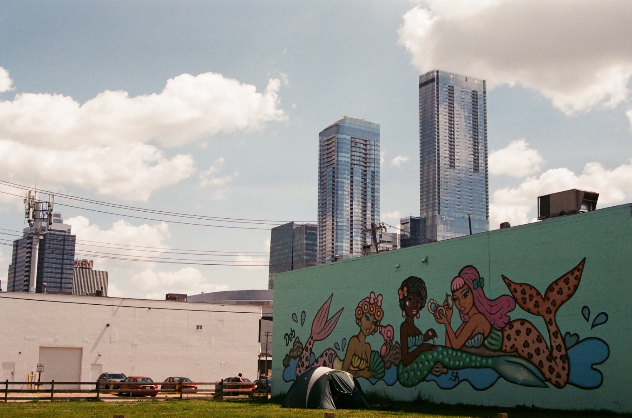 mural of three mermaids on a green wall with skyscrapers and a tent in the foreground.