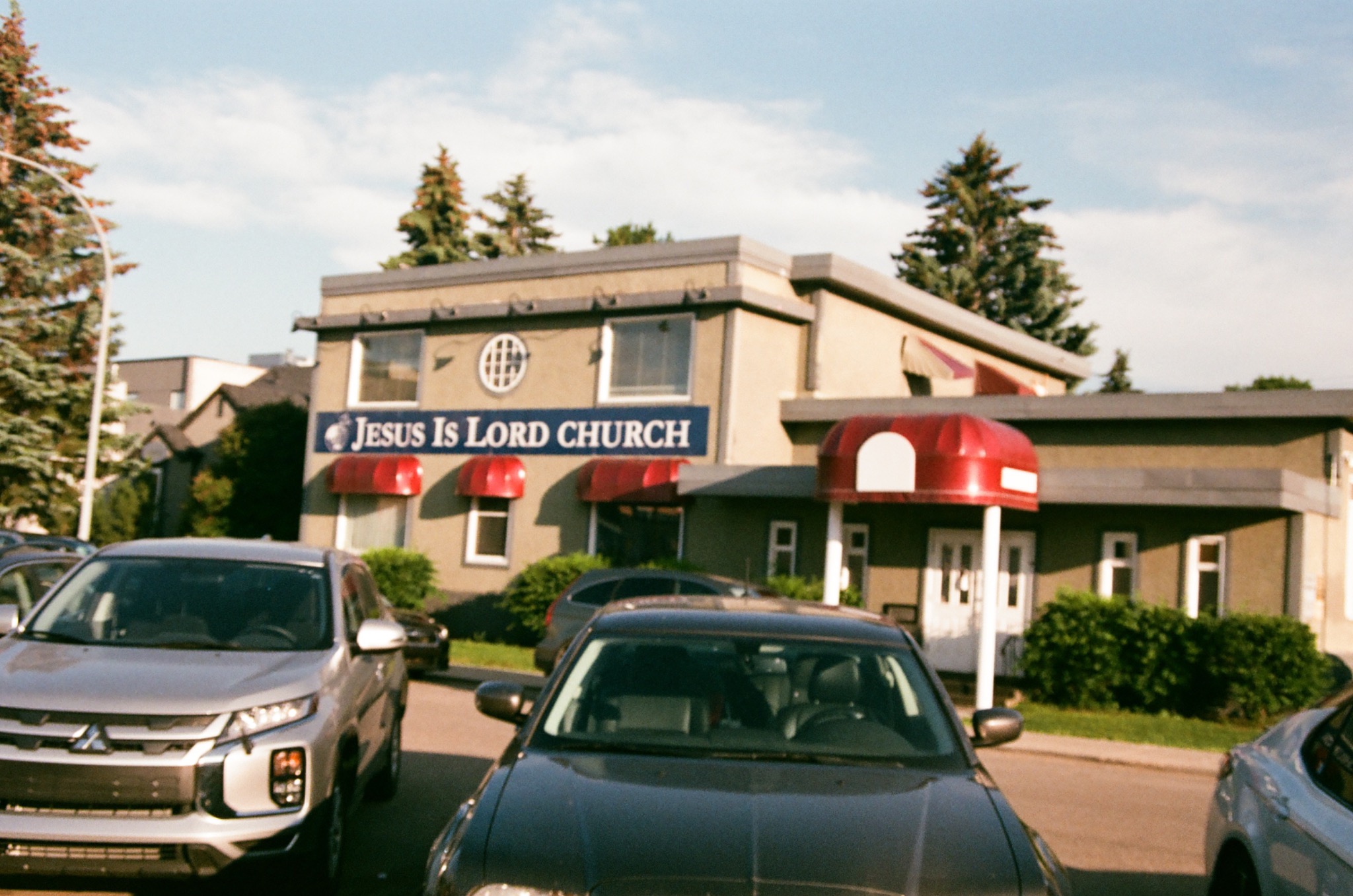 a beige church building with a blue 'jesus is lord church' sign, red awnings, and parked cars.